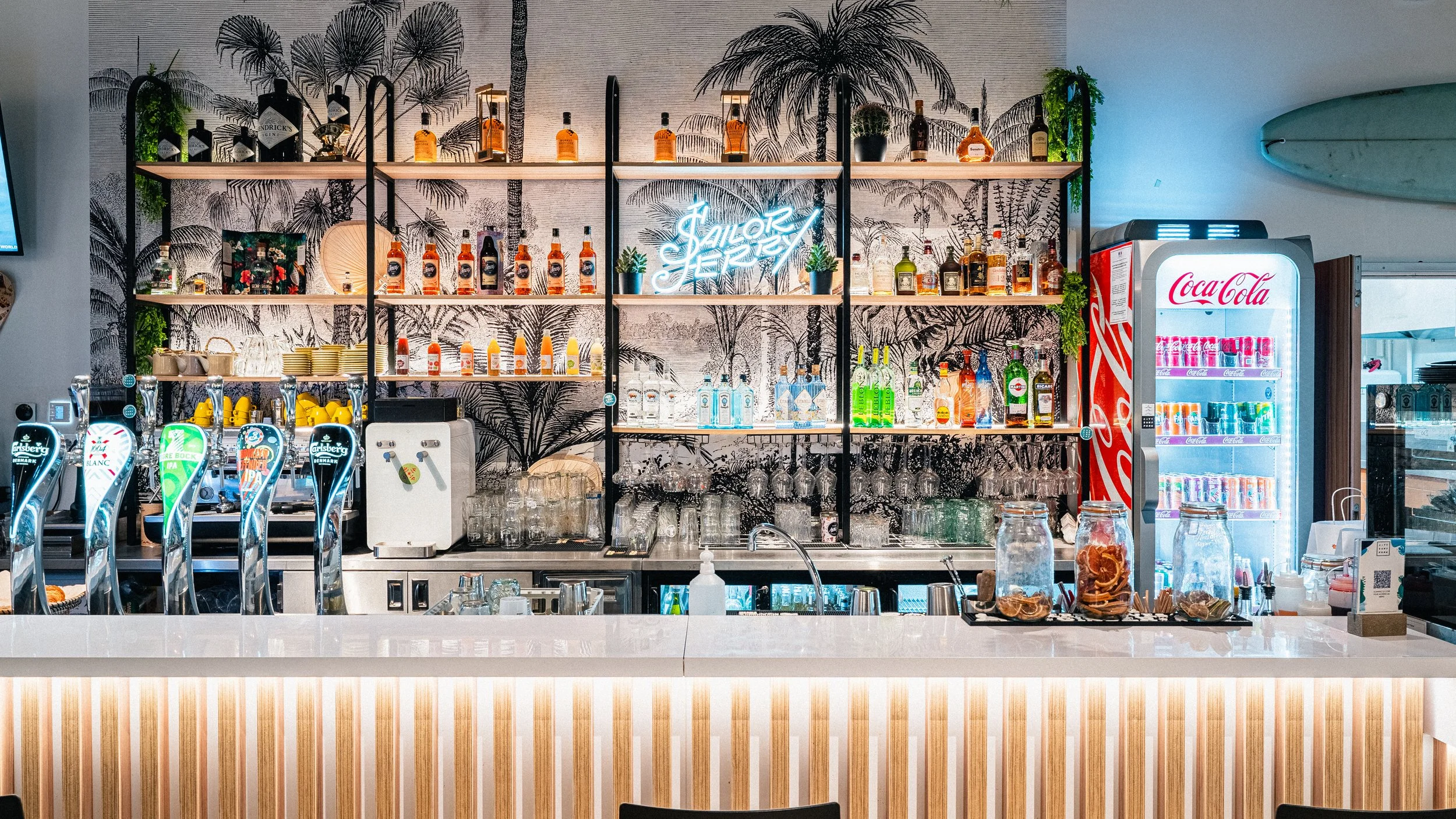 Bar avec étagères contenant des bouteilles d'alcool, un réfrigérateur Coca-Cola, et un comptoir avec des jars de fruits séchés, dans un décor tropical.